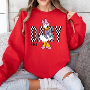 May include: Red sweatshirt with a checkered design and the word "DAISY" in white letters. A cartoon image of Daisy Duck is in the center of the design. The year "1940" is printed below the design.