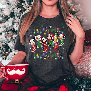 May include: A dark gray t-shirt with a Christmas design featuring Mickey Mouse, Donald Duck, Goofy, and Pluto wearing Christmas stockings. The design includes a string of Christmas lights and snowflakes.