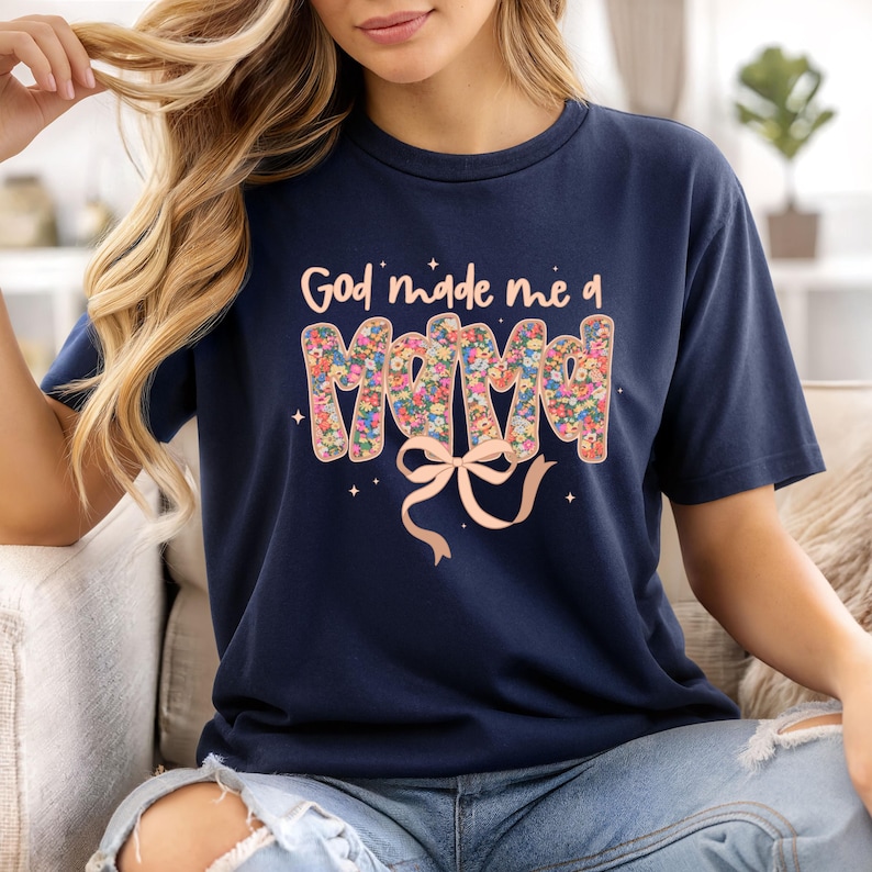 Pu&ograve; includere: T-shirt blu navy con la scritta "God made me a Mama". La parola "Mama" &egrave; riempita con un motivo floreale. Un fiocco rosa chiaro &egrave; sotto il testo, con piccoli accenti di stelle.