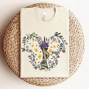 May include: A cream-colored t-shirt with a graphic of a purple and yellow tower with a green vine and flowers in the shape of Mickey Mouse ears.