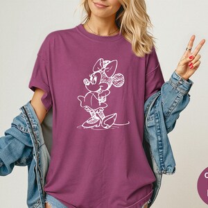May include: Berry-colored t-shirt featuring a white sketch of Minnie Mouse. The shirt has short sleeves and a relaxed fit. The image also shows a denim jacket. The text on the image says "Comfort Colors Berry".