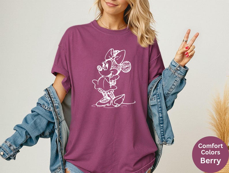 May include: Berry-colored t-shirt featuring a white sketch of Minnie Mouse. The shirt has short sleeves and a relaxed fit. The image also shows a denim jacket. The text on the image says "Comfort Colors Berry".