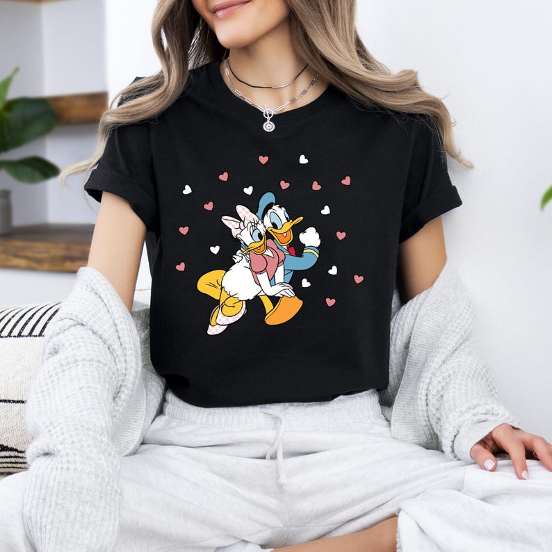 May include: Black t-shirt with a graphic print of Donald Duck and Daisy Duck holding each other. The graphic is surrounded by pink and white hearts.