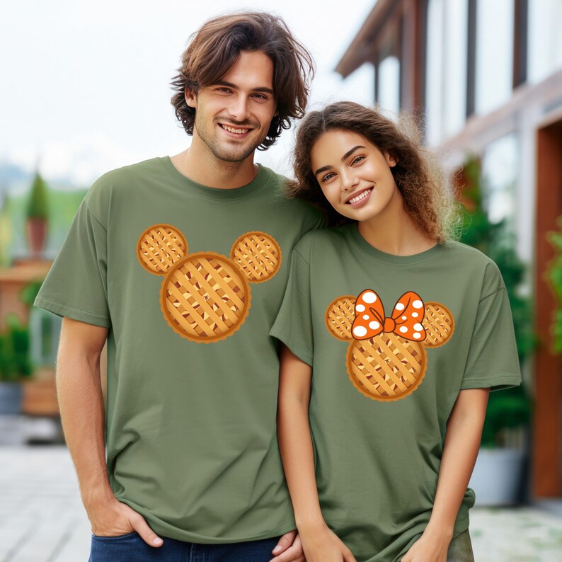 K&ouml;nnte beinhalten: Zwei Personen tragen olivgr&uuml;ne T-Shirts. Ein Shirt zeigt ein Mickey Mouse-Design in Kuchenform, das andere ein Minnie Mouse-Design in Kuchenform mit einer orangefarbenen Schleife mit wei&szlig;en Punkten. Die Shirts sind kurz&auml;rmlig.