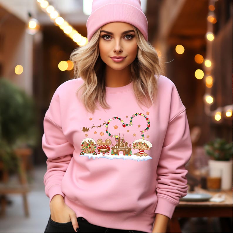 May include: Pink Christmas sweatshirt featuring a gingerbread Mickey Mouse design. The design includes a gingerbread Mickey Mouse head with a string of lights, a gingerbread castle, and gingerbread trees. The sweatshirt has a crew neck and long sleeves.