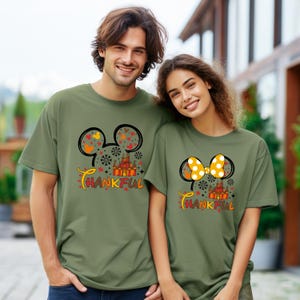 Puede incluir: Dos camisetas verde oliva con un dise&ntilde;o inspirado en Mickey Mouse. Una presenta la cabeza de Mickey con corazones y hojas, la otra con un lazo amarillo con lunares. Ambas camisetas tienen un castillo y la palabra "THANKFUL".