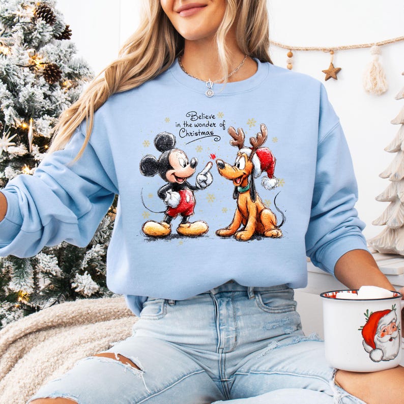 May include: Light blue sweatshirt featuring Mickey Mouse and Pluto in a Christmas design. Mickey is wearing red shorts and Pluto has a Santa hat and reindeer antlers. The text "Believe in the wonder of Christmas" is above the characters.