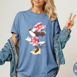 K&ouml;nnte beinhalten: Blaues Jeans-T-Shirt mit Minnie Mouse-Grafik. Die Zeichentrickfigur wird beim Auftragen von Lippenstift dargestellt und tr&auml;gt ein rosa gepunktetes Kleid und eine Schleife. Das Shirt hat kurze &Auml;rmel und eine lockere Passform. Die Worte "Minnie Mouse" sind kursiv geschrieben.