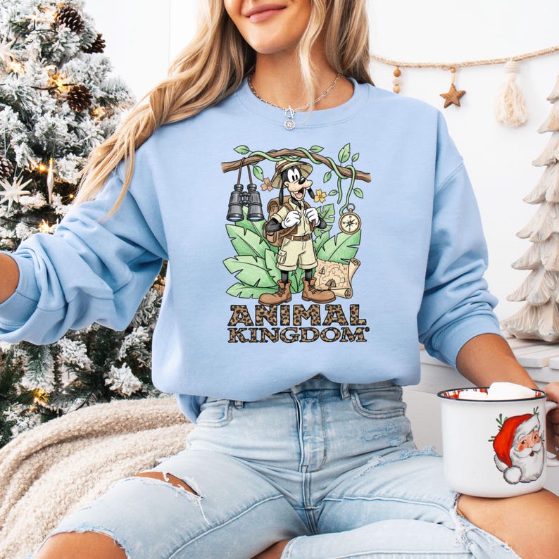 May include: Light blue sweatshirt featuring a cartoon graphic of Goofy in safari attire, with binoculars, a map, and the text "ANIMAL KINGDOM". The design is set against a backdrop of green leaves and branches.