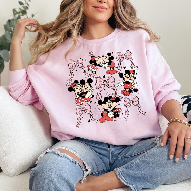 May include: Pink sweatshirt with a graphic print of Mickey Mouse and Minnie Mouse in various poses. The print features pink bows and hearts.
