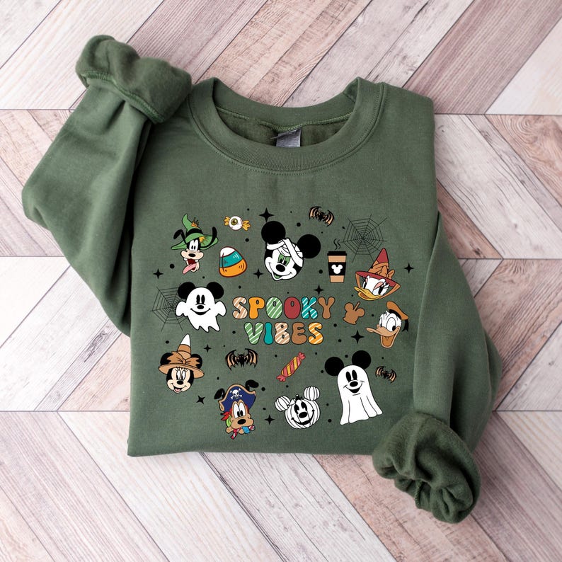 May include: A green sweatshirt with a Halloween-themed design featuring Mickey Mouse and friends. The design includes a ghost, a spiderweb, candy corn, and the text "Spooky Vibes".