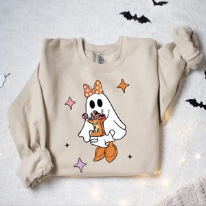 May include: Beige sweatshirt featuring a cartoon ghost with a polka-dot bow, holding a "Trick or Treat" bag filled with candy. The ghost is wearing orange shoes and surrounded by star shapes. Halloween-themed apparel.