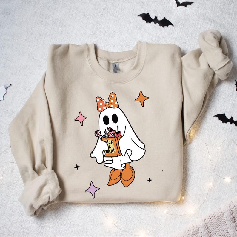 May include: Beige sweatshirt featuring a cartoon ghost with a polka-dot bow, holding a "Trick or Treat" bag filled with candy. The ghost is wearing orange shoes and surrounded by star shapes. Halloween-themed apparel.