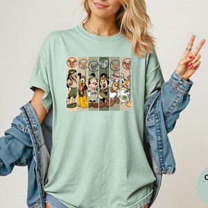 May include: A light green t-shirt featuring a graphic of cartoon characters in safari outfits. The characters include Mickey Mouse, Donald Duck, and Goofy. The shirt is a Comfort Colors Bay.