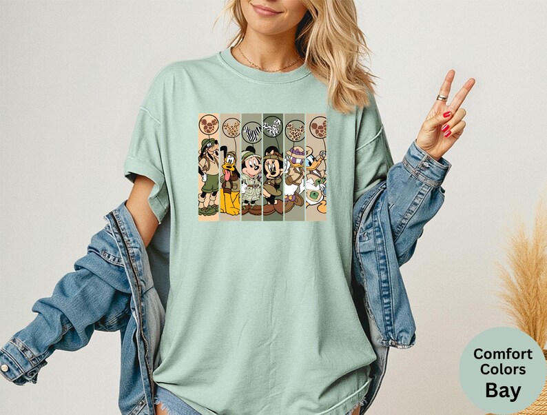 May include: A light green t-shirt featuring a graphic of cartoon characters in safari outfits. The characters include Mickey Mouse, Donald Duck, and Goofy. The shirt is a Comfort Colors Bay.