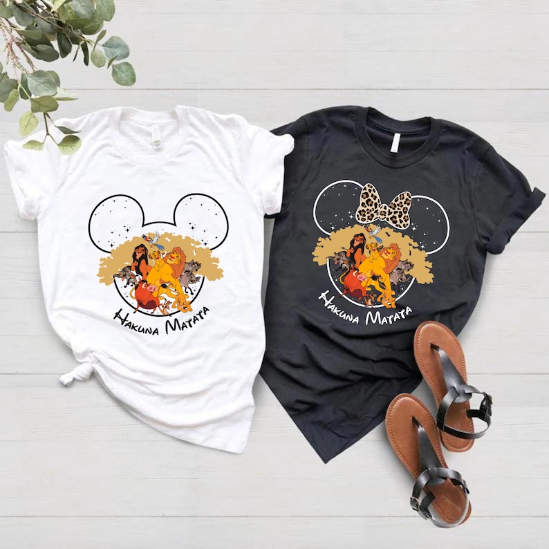 Puede incluir: Dos camisetas con un dise&ntilde;o de cabeza de Mickey Mouse en blanco y negro con los personajes de El Rey Le&oacute;n Simba, Nala, Tim&oacute;n y Pumba. El texto "Hakuna Matata" est&aacute; impreso debajo del dise&ntilde;o. Una camiseta es blanca y la otra es negra.