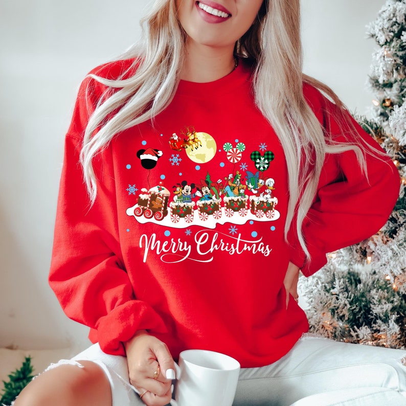 May include: Red Christmas sweatshirt with a graphic of Mickey Mouse and friends riding a gingerbread train. The text "Merry Christmas" is printed below the train.
