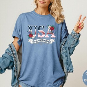 May include: A blue jean-colored t-shirt with the word "USA" in a patriotic design, incorporating the American flag and floral accents. The shirt also features the text "250 years of freedom" on a banner.