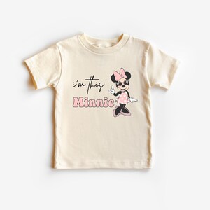 Pu&ograve; includere: T-shirt color crema per bambini piccoli con grafica di Minnie Mouse. La grafica include il testo "I'm this Minnie" in nero e rosa, insieme a un personaggio dei cartoni animati di Minnie Mouse con occhiali da sole e un vestito a pois.