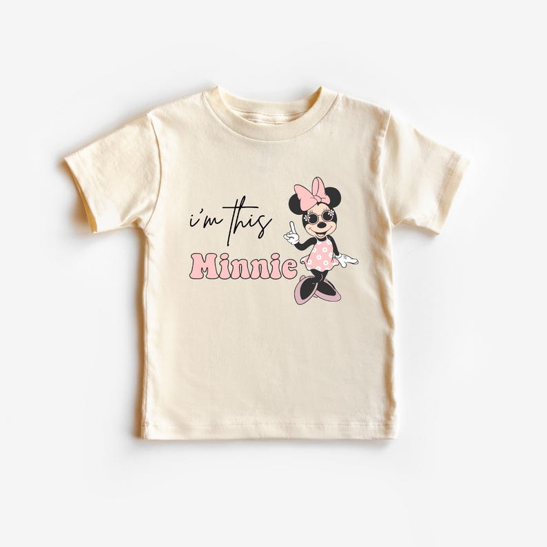 Pu&ograve; includere: T-shirt color crema per bambini piccoli con grafica di Minnie Mouse. La grafica include il testo "I'm this Minnie" in nero e rosa, insieme a un personaggio dei cartoni animati di Minnie Mouse con occhiali da sole e un vestito a pois.