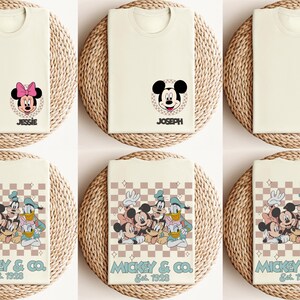 May include: Six white t-shirts with different Disney character designs. The shirts feature Mickey Mouse, Minnie Mouse, Donald Duck, Goofy, and Pluto. The shirts have a checkered pattern and the text "Mickey & Co. Est. 1928".