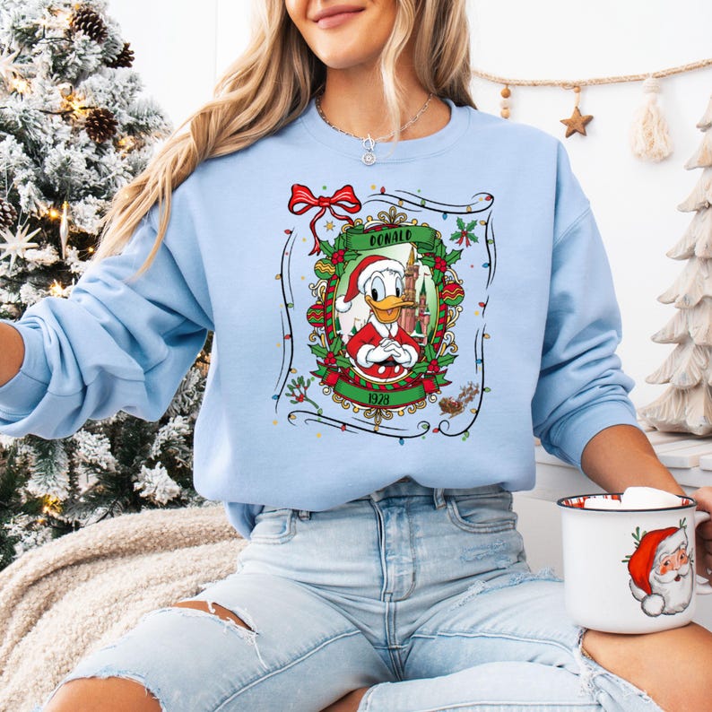 Może przedstawiać: Jasnoniebieska bluza z świąteczną grafiką Kaczora Donalda w czapce Świętego Mikołaja, z napisem "DONALD" i "1928". Projekt jest obramowany ostrokrzewem, światłami i czerwoną kokardą. Widoczny jest r&oacute;wnież kubek o tematyce świątecznej.