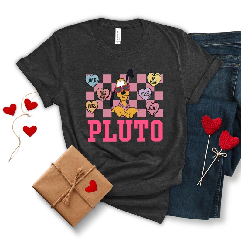 May include: Dark gray t-shirt featuring Pluto wearing sunglasses, surrounded by heart-shaped candies with phrases like "Kisses" and "Be Mine." The word "PLUTO" is printed in pink block letters. A pair of blue jeans and Valentine's Day decorations are also visible.
