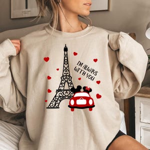 May include: A beige sweatshirt with a graphic print of the Eiffel Tower, a red car with Mickey Mouse and Minnie Mouse figures inside, and the text "I'm always with you". The graphic is surrounded by red hearts.