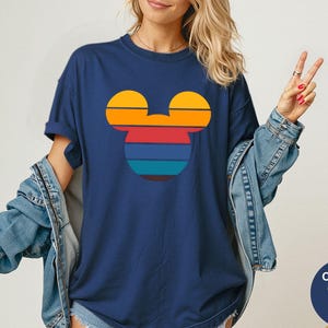 K&ouml;nnte beinhalten: Marineblaues T-Shirt mit einem Retro-Mickey-Mouse-Silhouette-Design in Sonnenuntergangsfarben. Die Grafik enth&auml;lt horizontale Streifen in Orange, Gelb, Rot und Blau. Das Shirt ist im Comfort Colors-Stil.
