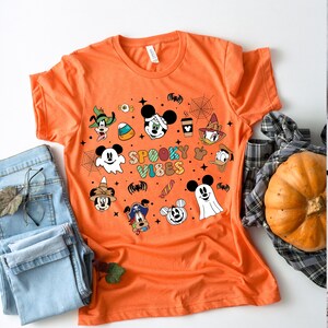 May include: Orange t-shirt with a Halloween theme featuring Mickey Mouse and friends. The shirt has a graphic print of Mickey Mouse, Minnie Mouse, Donald Duck, Goofy, and Pluto dressed in Halloween costumes. The text "Spooky Vibes" is printed on the shirt.