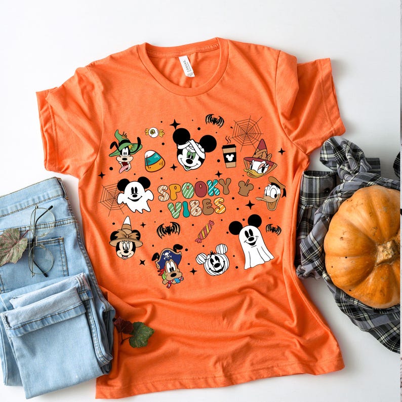 May include: Orange t-shirt with a Halloween theme featuring Mickey Mouse and friends. The shirt has a graphic print of Mickey Mouse, Minnie Mouse, Donald Duck, Goofy, and Pluto dressed in Halloween costumes. The text "Spooky Vibes" is printed on the shirt.