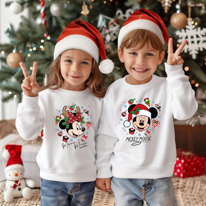 May include: Two children wearing white sweatshirts and Santa hats pose in front of a Christmas tree. The sweatshirts feature Mickey and Minnie Mouse in festive holiday designs. One says "Minnie Mouse Forever" and the other says "Mickey Mouse".