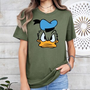May include: Olive green t-shirt featuring a cartoon graphic of Donald Duck's face. The graphic includes a blue sailor hat, black hair, and an orange beak. The t-shirt is a crew neck style.