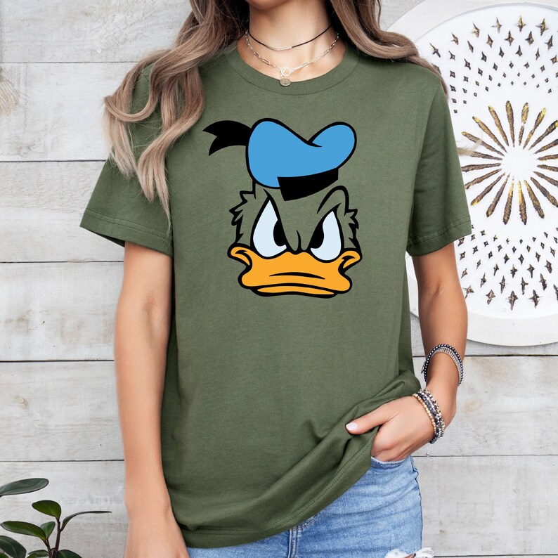 May include: Olive green t-shirt featuring a cartoon graphic of Donald Duck's face. The graphic includes a blue sailor hat, black hair, and an orange beak. The t-shirt is a crew neck style.