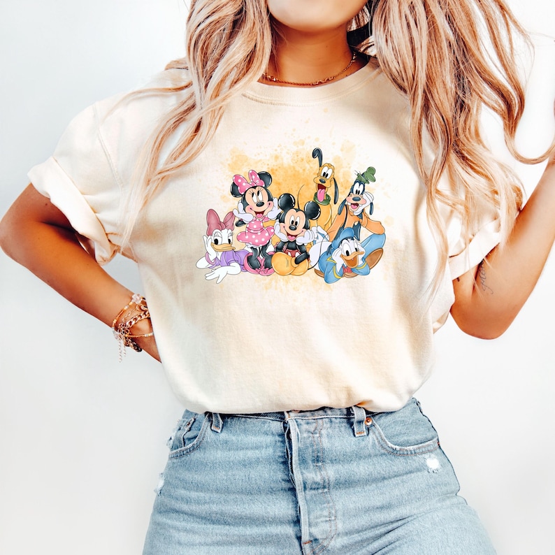 Puede incluir: Una camiseta blanca con un aspecto amarillento y descolorido presenta una imagen de dibujos animados de Mickey Mouse, Minnie Mouse, Donald Duck, Goofy y Pluto. Los personajes sonr&iacute;en y parecen estar sentados juntos.