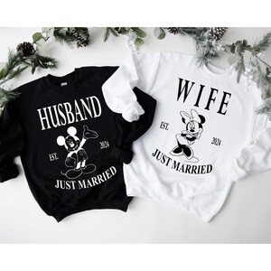 May include: Black and white sweatshirt with the text "Husband" and a Mickey Mouse illustration. White text reads "Est. 2024 Just Married".  White sweatshirt with the text "Wife" and a Minnie Mouse illustration. White text reads "Est. 2024 Just Married".