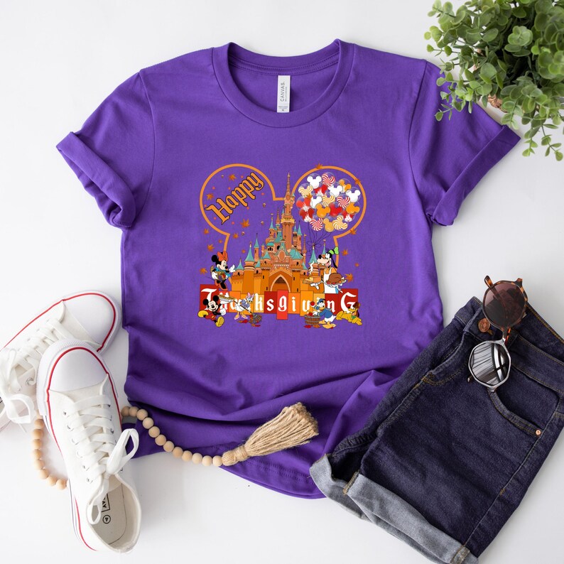 May include: A purple t-shirt featuring a graphic of a castle, Mickey Mouse, and other Disney characters. The design includes the words "Happy Thanksgiving" in a circular Mickey Mouse ear shape. White sneakers and denim shorts are also in the image.