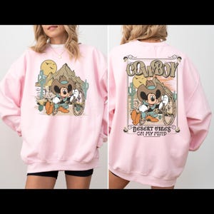 May include: Pink sweatshirt with a cartoon Mickey Mouse wearing a cowboy hat and holding a lasso. The front of the sweatshirt has a desert scene with a mountain and cacti. The back of the sweatshirt says "COWBOY" in a vintage font and "DESERT VIBES ON MY MIND".