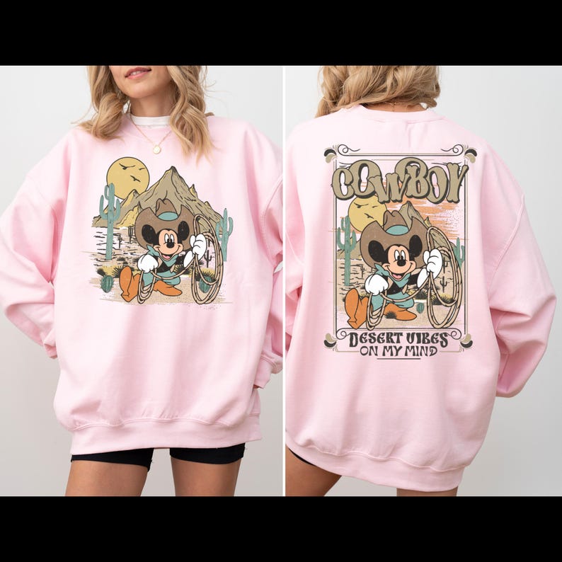 May include: Pink sweatshirt with a cartoon Mickey Mouse wearing a cowboy hat and holding a lasso. The front of the sweatshirt has a desert scene with a mountain and cacti. The back of the sweatshirt says "COWBOY" in a vintage font and "DESERT VIBES ON MY MIND".