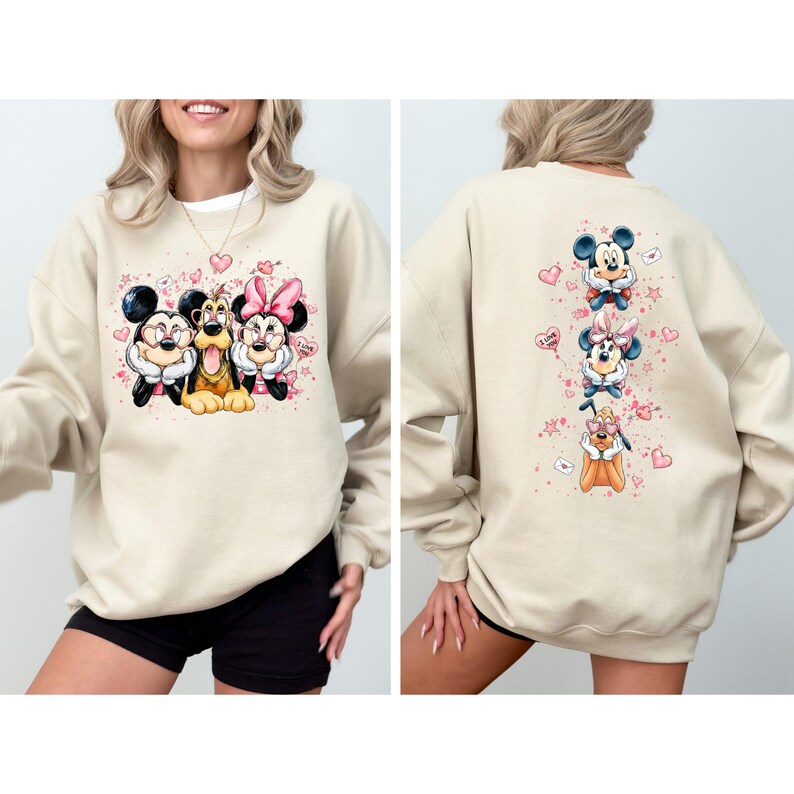 May include: Beige sweatshirt featuring Mickey Mouse, Goofy, and Minnie Mouse with pink hearts and stars. The back of the sweatshirt shows the same characters in a vertical design. A fun, casual top for Disney fans.