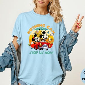 May include: Light blue t-shirt featuring Mickey and Minnie Mouse in a red car, with the text "NOTHING CAN STOP US NOW!". The design is set against a colorful sunset backdrop. The shirt is made of Comfort Colors Chambray.
