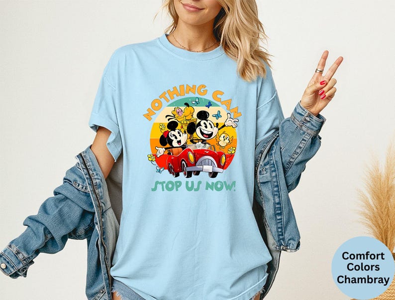 May include: Light blue t-shirt featuring Mickey and Minnie Mouse in a red car, with the text "NOTHING CAN STOP US NOW!". The design is set against a colorful sunset backdrop. The shirt is made of Comfort Colors Chambray.