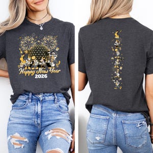 May include: Dark gray t-shirt with a festive "Happy New Year 2026" design featuring fireworks and cartoon characters. The back of the shirt has a vertical design of the same characters. The shirt is paired with blue jeans.