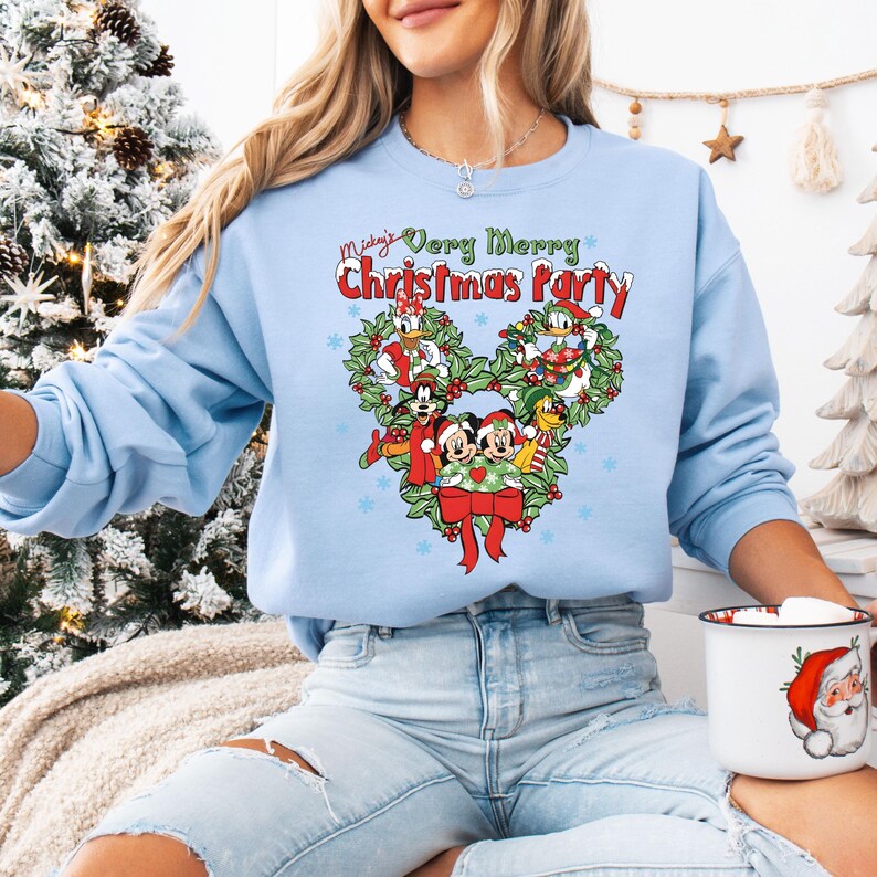 Op de afbeelding: Een lichtblauwe sweatshirt met een Mickey Mouse-thema kerstontwerp. De afbeelding bevat Mickey, Minnie, Donald en Daisy Duck, omringd door een krans en de tekst "Mickey's Very Merry Christmas Party."