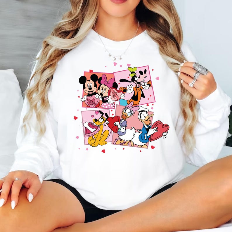 May include: White sweatshirt with a pink and white graphic print of Mickey Mouse, Minnie Mouse, Goofy, Donald Duck, and Pluto holding hearts. The characters are surrounded by pink hearts.