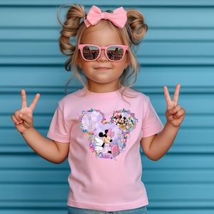 May include: A pink t-shirt with a floral design featuring a cartoon image of Minnie Mouse with wings and other Disney characters. The shirt has short sleeves and a crew neck.
