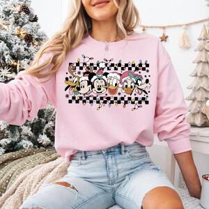 May include: Pink Christmas themed sweatshirt with a checkered pattern and a row of Disney characters including Mickey Mouse, Minnie Mouse, Donald Duck, Daisy Duck, and Goofy.
