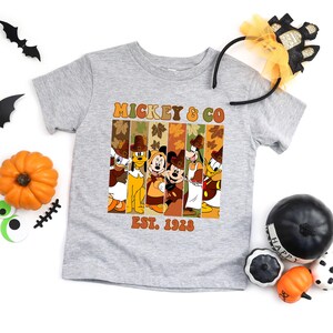 May include: A gray t-shirt featuring a graphic of Mickey Mouse and friends in fall attire. The graphic includes the text "MICKEY & CO" and "EST. 1928". Halloween decorations, including pumpkins and bats, are arranged around the shirt.