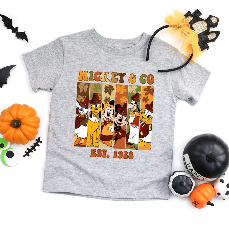 May include: A gray t-shirt featuring a graphic of Mickey Mouse and friends in fall attire. The graphic includes the text "MICKEY & CO" and "EST. 1928". Halloween decorations, including pumpkins and bats, are arranged around the shirt.