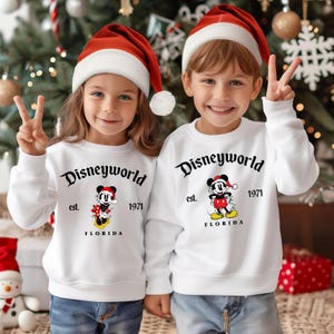 May include: Two children wearing white sweatshirts with a Mickey Mouse design and the text "Disneyworld est. 1971 Florida". The children are wearing red Santa hats and smiling.